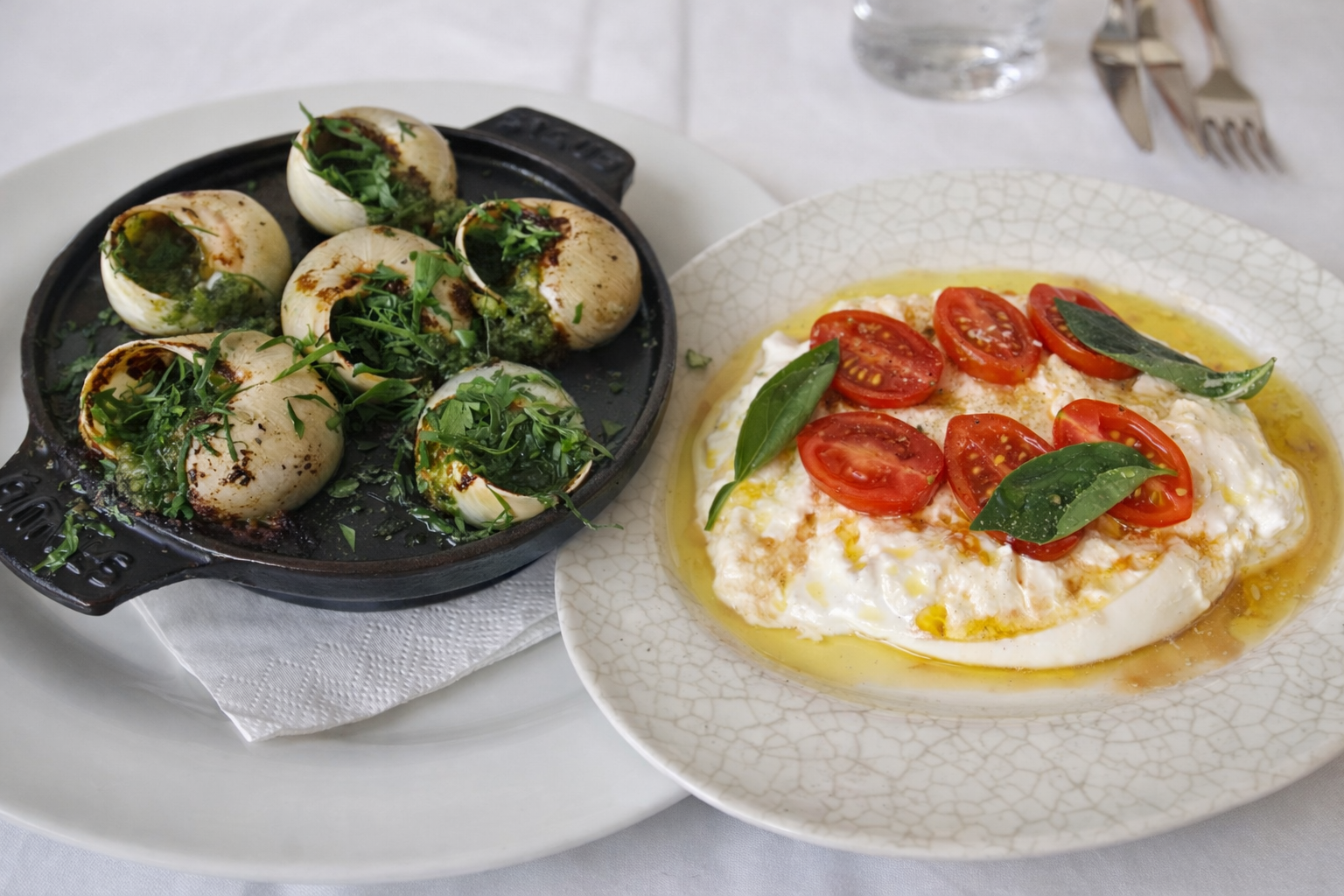 Burrata with cherry tomatoes and basil served next to garlic herb escargot in a cast-iron pan on a white table setting.
